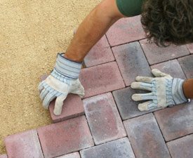 A top-down view of a man installing red and black paving stones on a bed of sand.