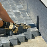 Gloved hands laying paving stones on a bed of compact bedding sand.